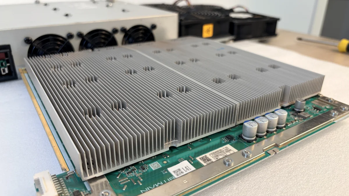 Placa hash de un Antminer T21 con el disipador hacia arriba: bloque de aletas a lo largo de toda la placa, fila de condensadores y puntos de fijación, al fondo la fuente y los ventiladores