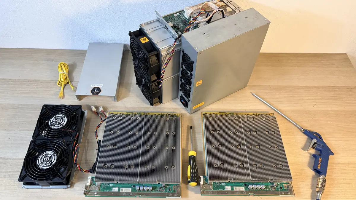 Antminer T21 desmontado sobre el banco: carcasa, tapa de la placa de control, fuente de alimentación, dos bloques de ventiladores, dos placas hash con los disipadores hacia arriba y una pistola de aire comprimido